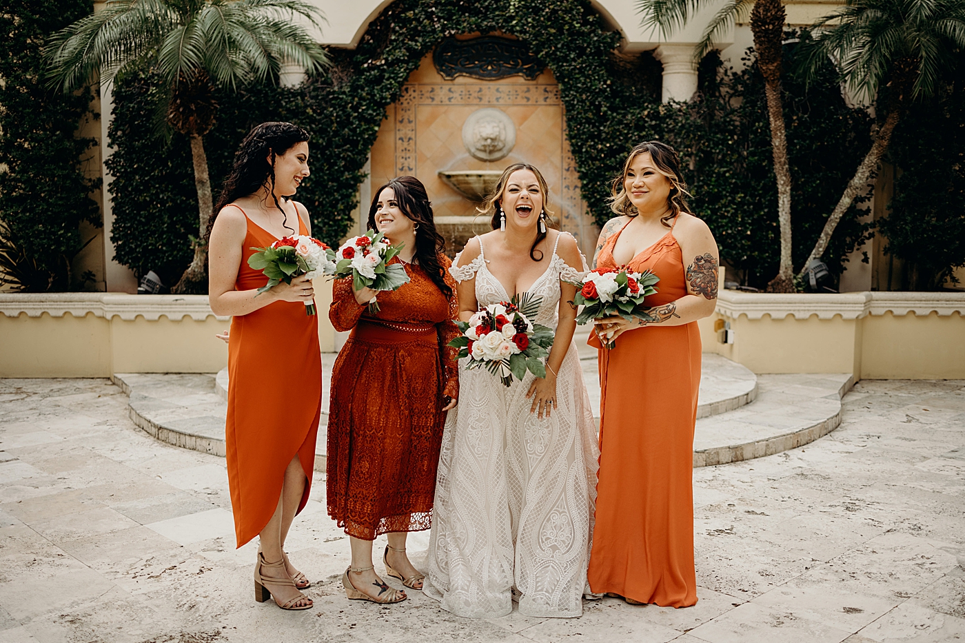 Bride and sunset orange dressed Bridesmaids in front of fountain with red and white floral bouquets Benvenuto Restaurant Wedding Photography captured by South Florida Wedding Photographer Maggie Alvarez Photography
