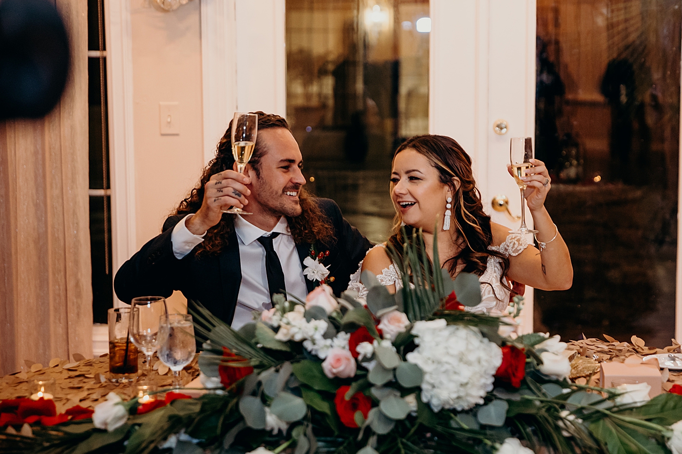 Bride and Groom holding up Champaign glasses for cheers at sweetheart table Benvenuto Restaurant Wedding Photography captured by South Florida Wedding Photographer Maggie Alvarez Photography