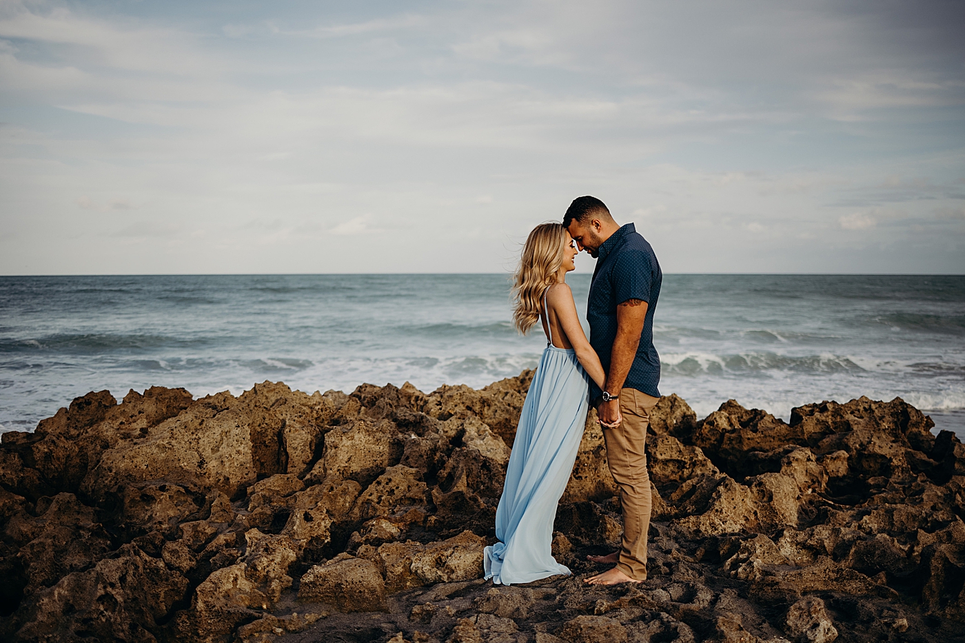 Couple holding hands and resting on each others heads with course rocks with ocean waves behind them Coral Cove Park Engagement Photography captured by Maggie Alvarez Photography