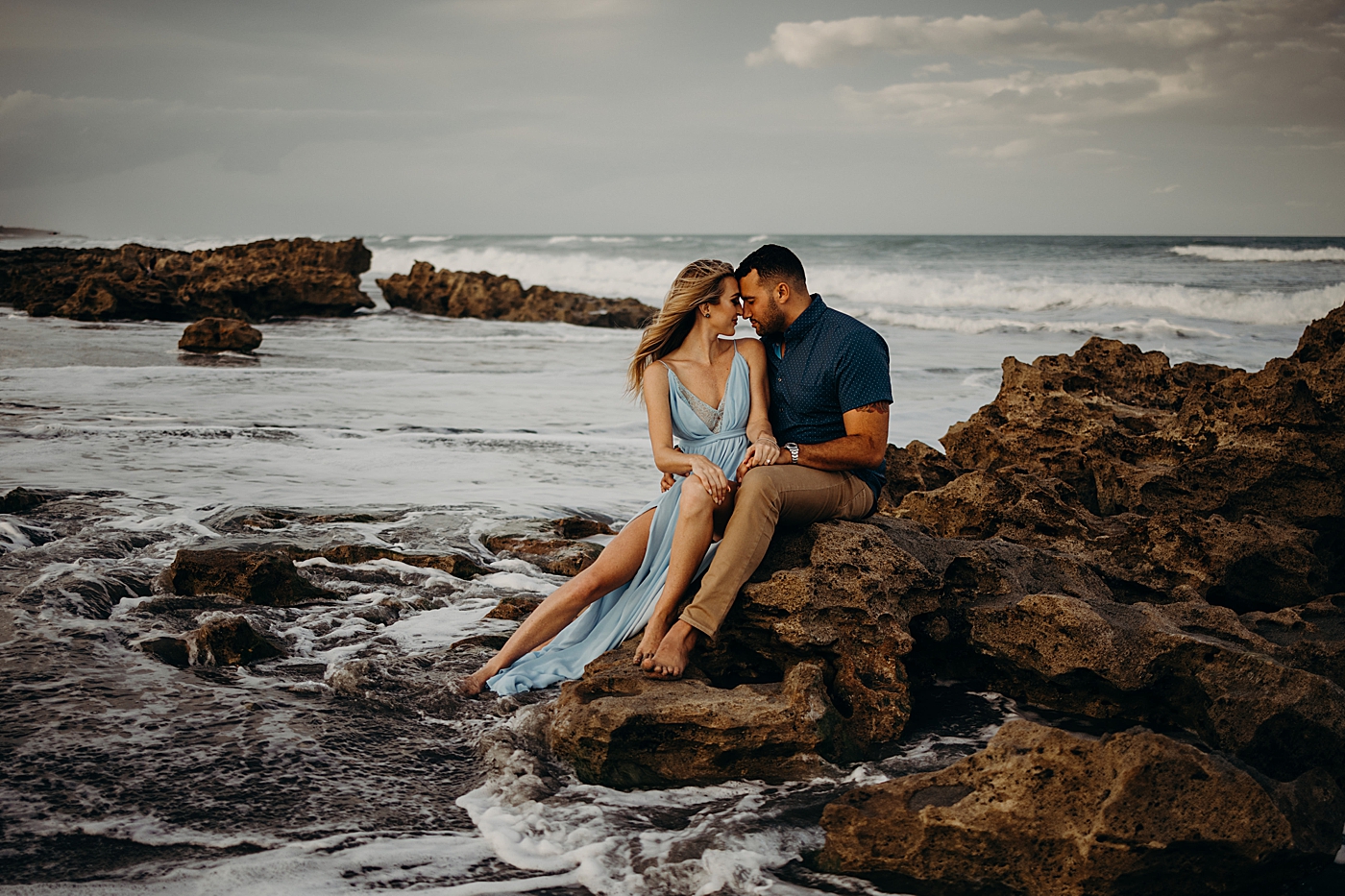 Couple sitting on the rock holding each other as the waves come in Coral Cove Park Engagement Photography captured by Maggie Alvarez Photography