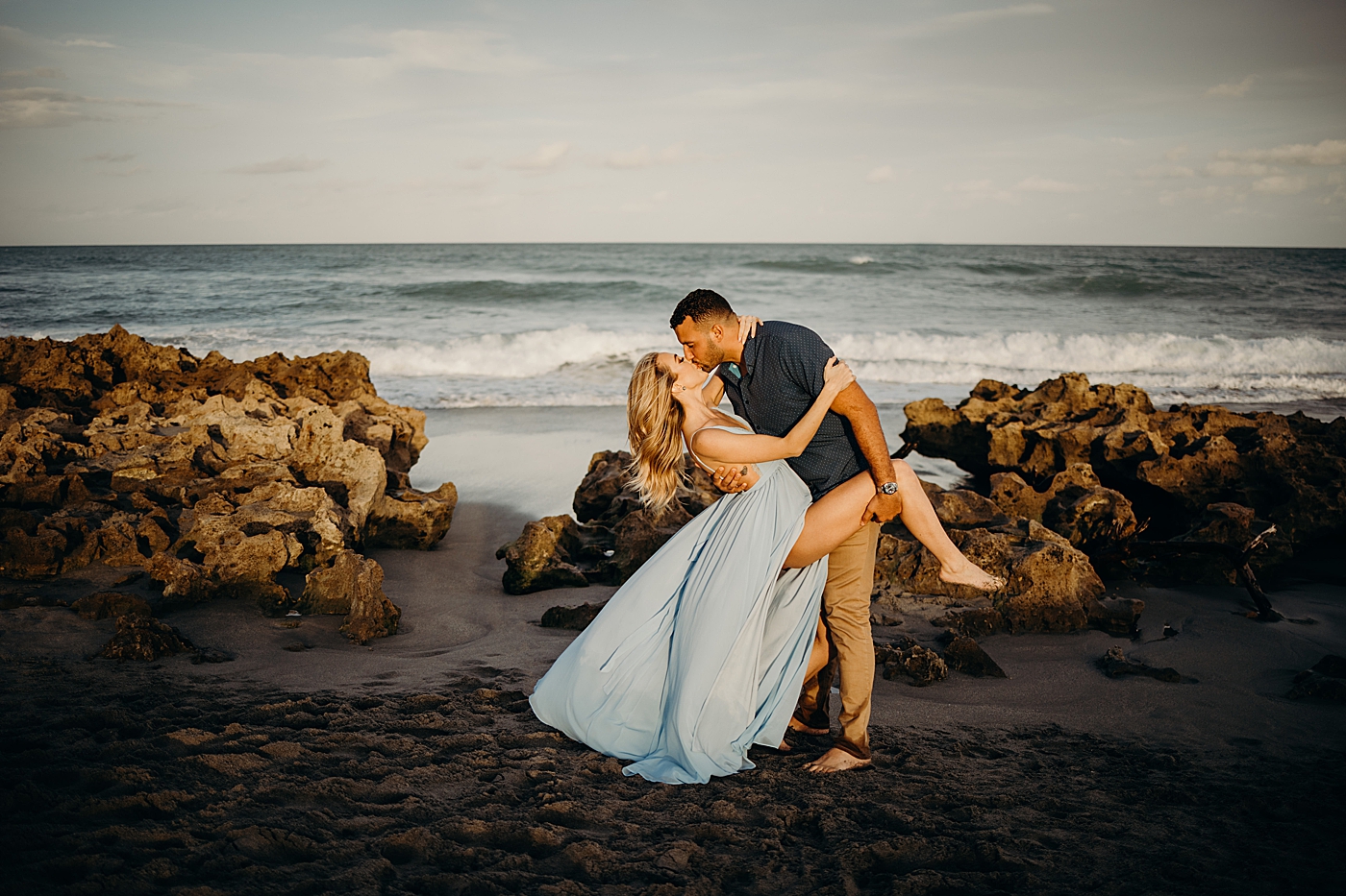 Couple dipping and kissing on beach as waves come in on rocky beach Coral Cove Park Engagement Photography captured by Maggie Alvarez Photography