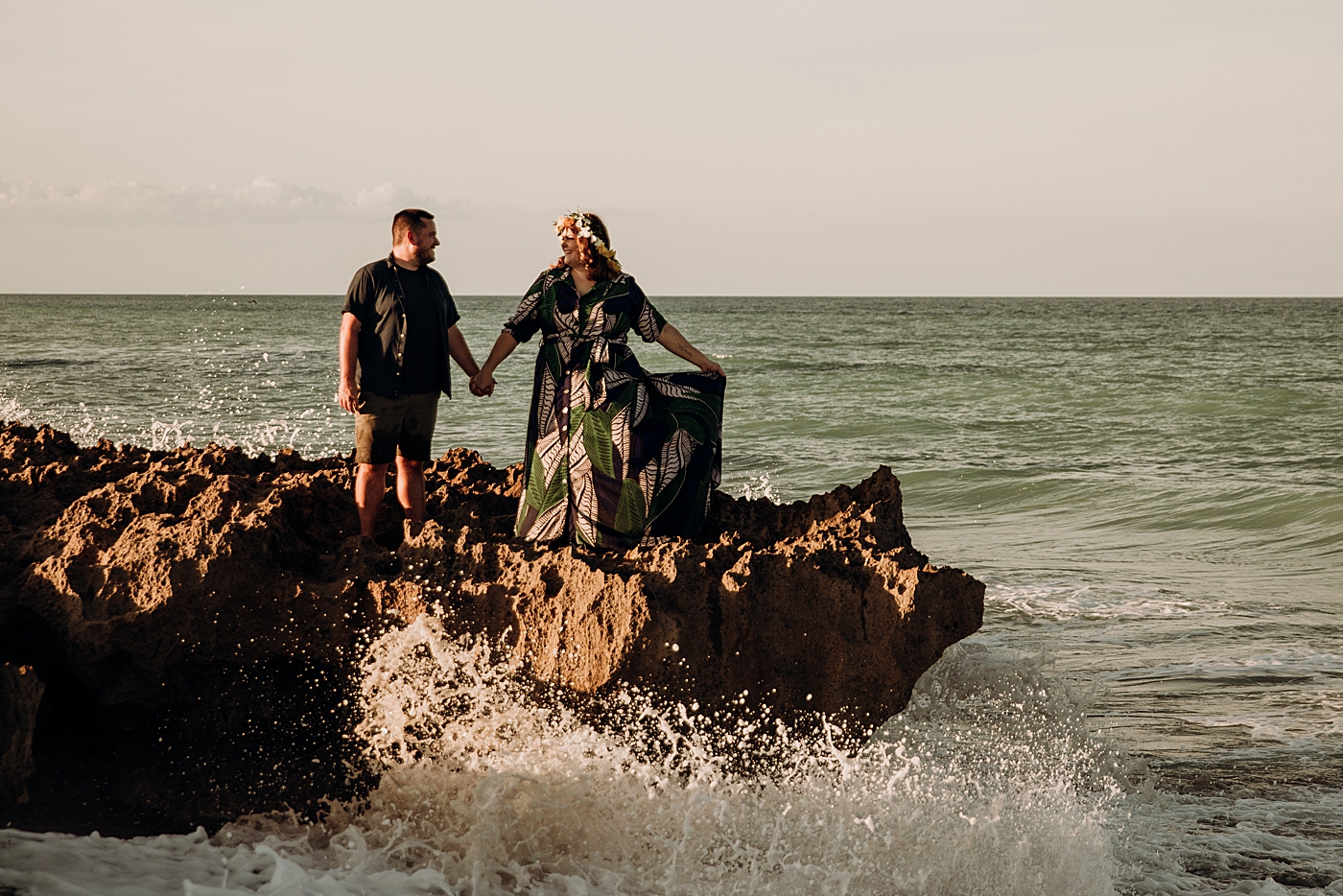 Couple standing on rock on the beach with crashing ocean wave House of Refuge Engagement Photography captured by Maggie Alvarez Photography