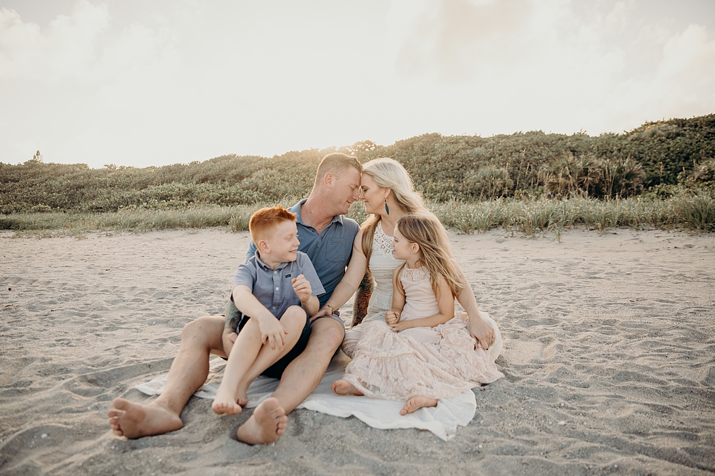 Family sitting on beach towel with parents nuzzling with the sun setting Ocean Ridge Hammock Park Family Photography by South Florida Family Photographer Maggie Alvarez Photography