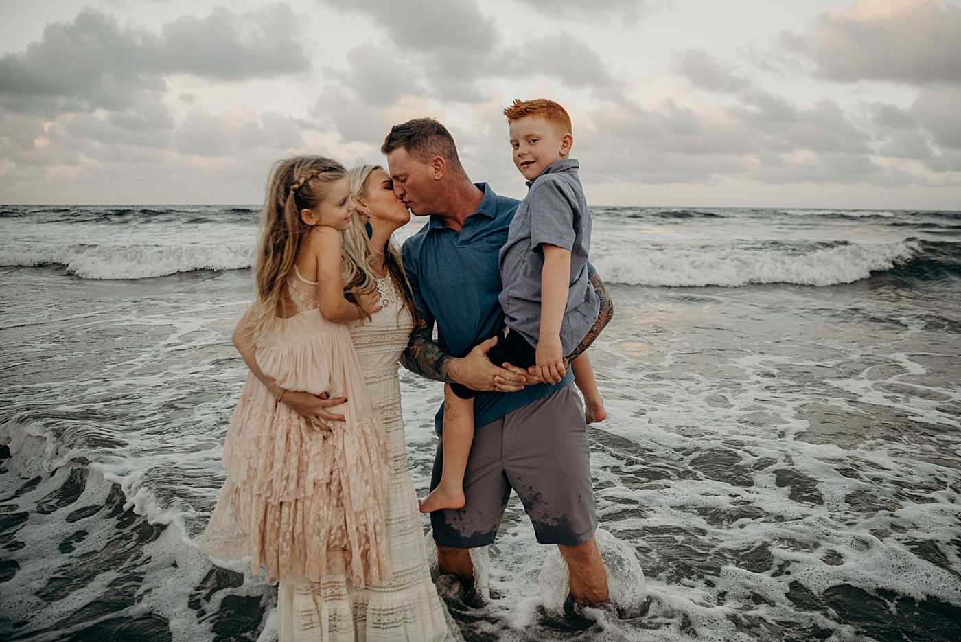 Parents standing in ocean water kissing each other and holding children in their arms Ocean Ridge Hammock Park Family Photography by South Florida Family Photographer Maggie Alvarez Photography
