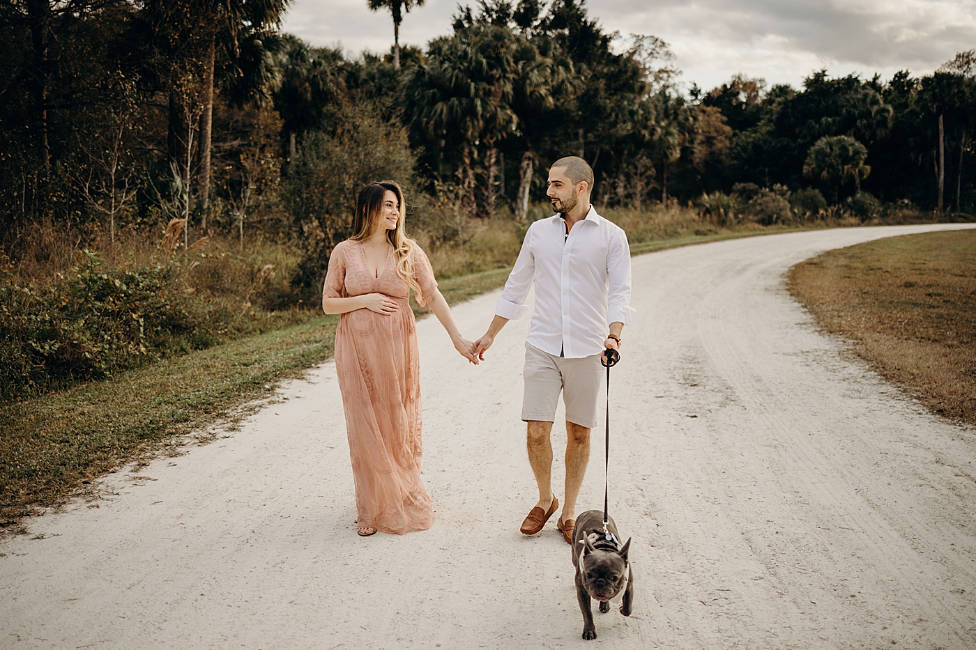 Couple holding hands and walking together on dirt path while walking dog Riverbend Park Maternity Photography by South Florida Family Photographer Maggie Alvarez Photography