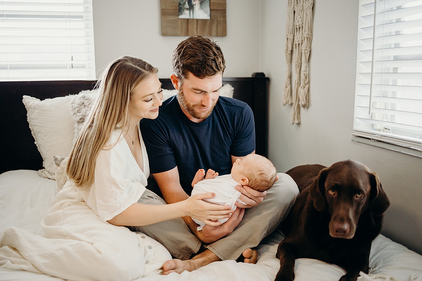 Parents looking at swaddled baby with dog next to them on the bed South Florida Newborn Photography captured by South Florida Family Photographer Maggie Alvarez Photography