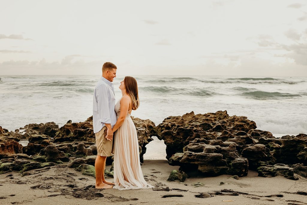 Couple holding hands and looking at each other in front of drift rocks on the beach Coral Cove Park Engagement Photography captured by South Florida Family Photographer Maggie Alvarez Photography