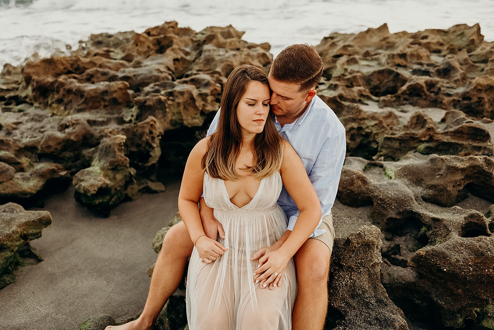 Couple sitting on drift rocks on the beach nuzzling Coral Cove Park Engagement Photography captured by South Florida Family Photographer Maggie Alvarez Photography