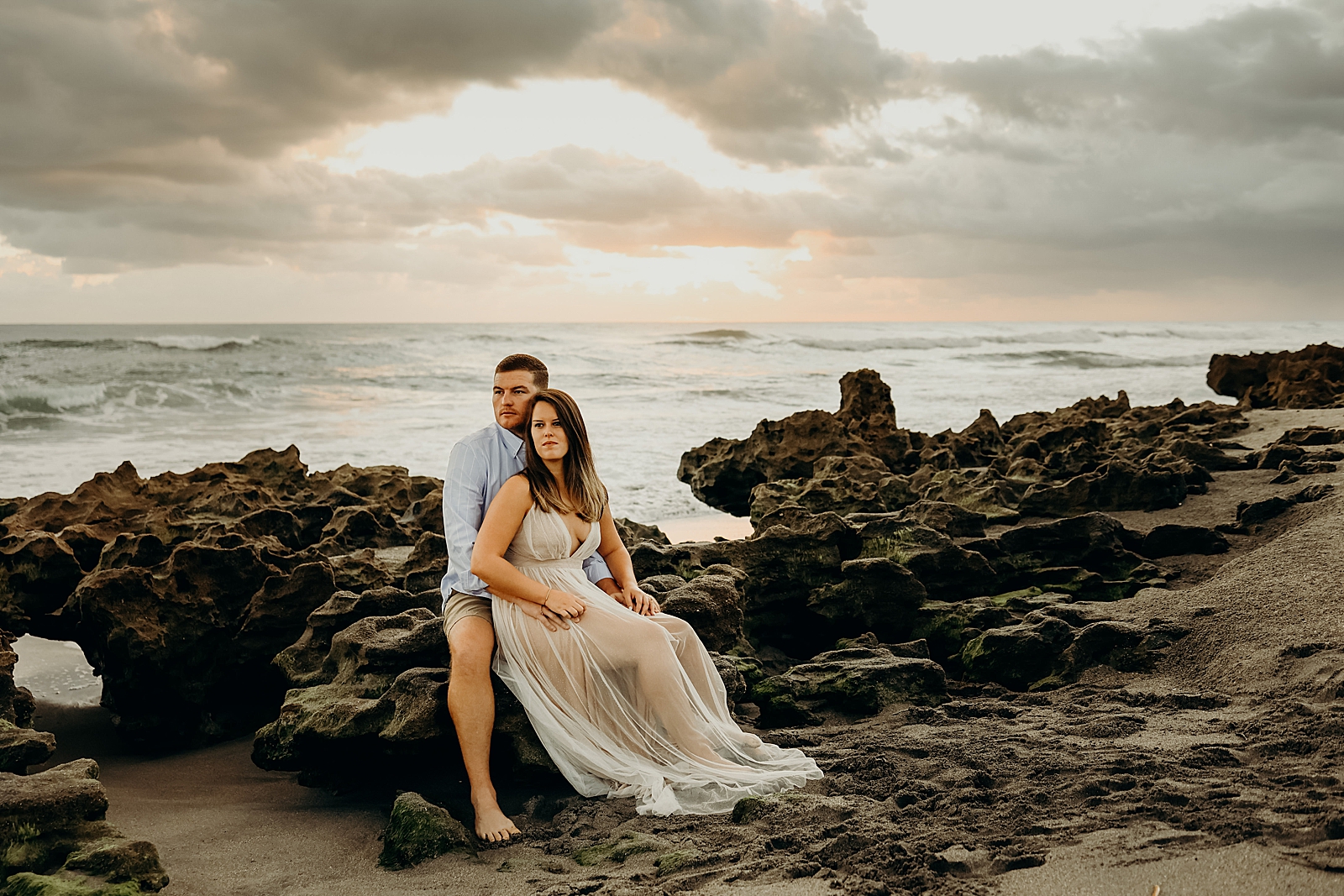 Man holding woman sitting on drift rock by the ocean Coral Cove Park Engagement Photography captured by South Florida Family Photographer Maggie Alvarez Photography