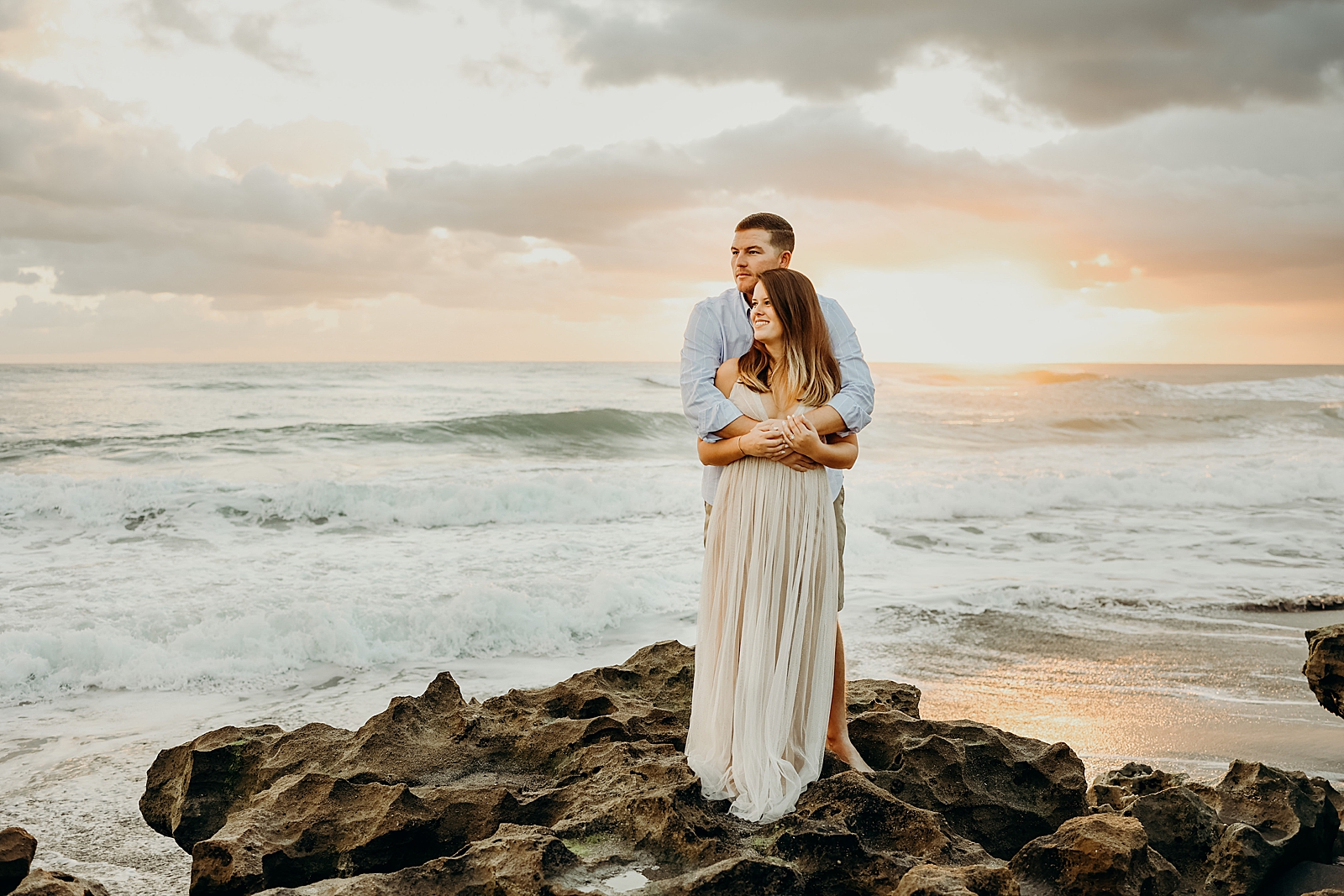 Couple holding each other standing on drift rock on the beach Coral Cove Park Engagement Photography captured by South Florida Family Photographer Maggie Alvarez Photography