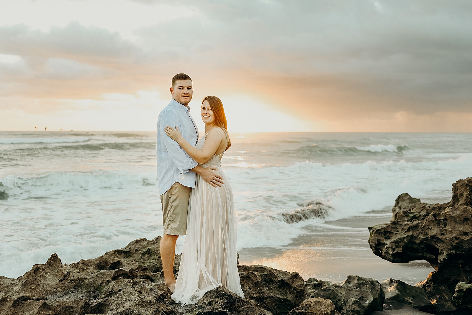 Couple holding each other looking at the camera on beach rock as waves come in Coral Cove Park Engagement Photography captured by South Florida Family Photographer Maggie Alvarez Photography