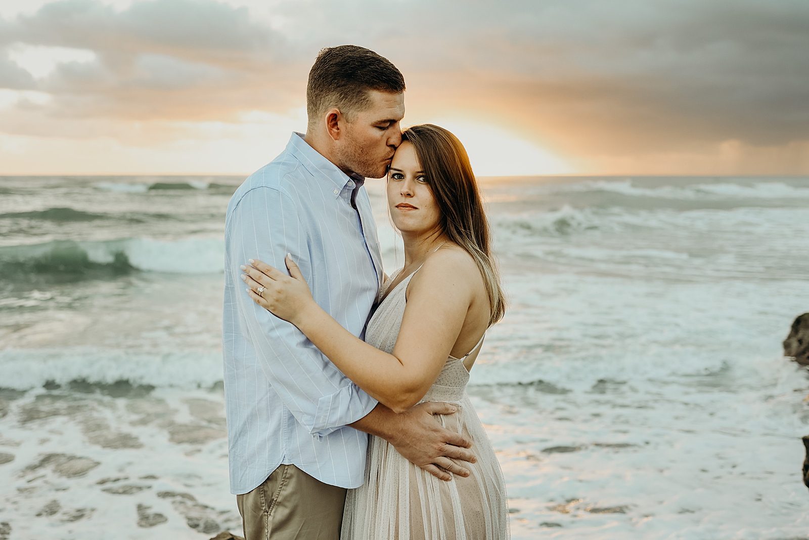 Man kisses woman on the side of her head in front of ocean waves Coral Cove Park Engagement Photography captured by South Florida Family Photographer Maggie Alvarez Photography