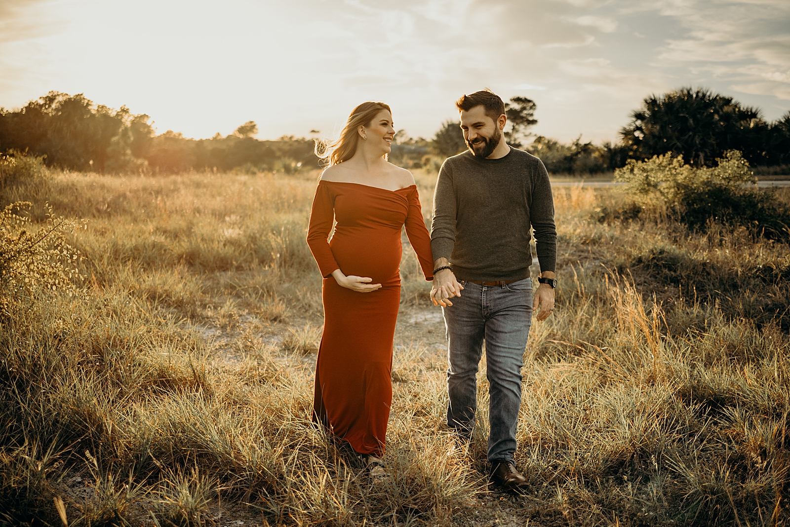Couple holding hands strolling on tall grass field Johnathan Dickinson Park Maternity Photography captured by South Florida Family Photographer Maggie Alvarez Photography