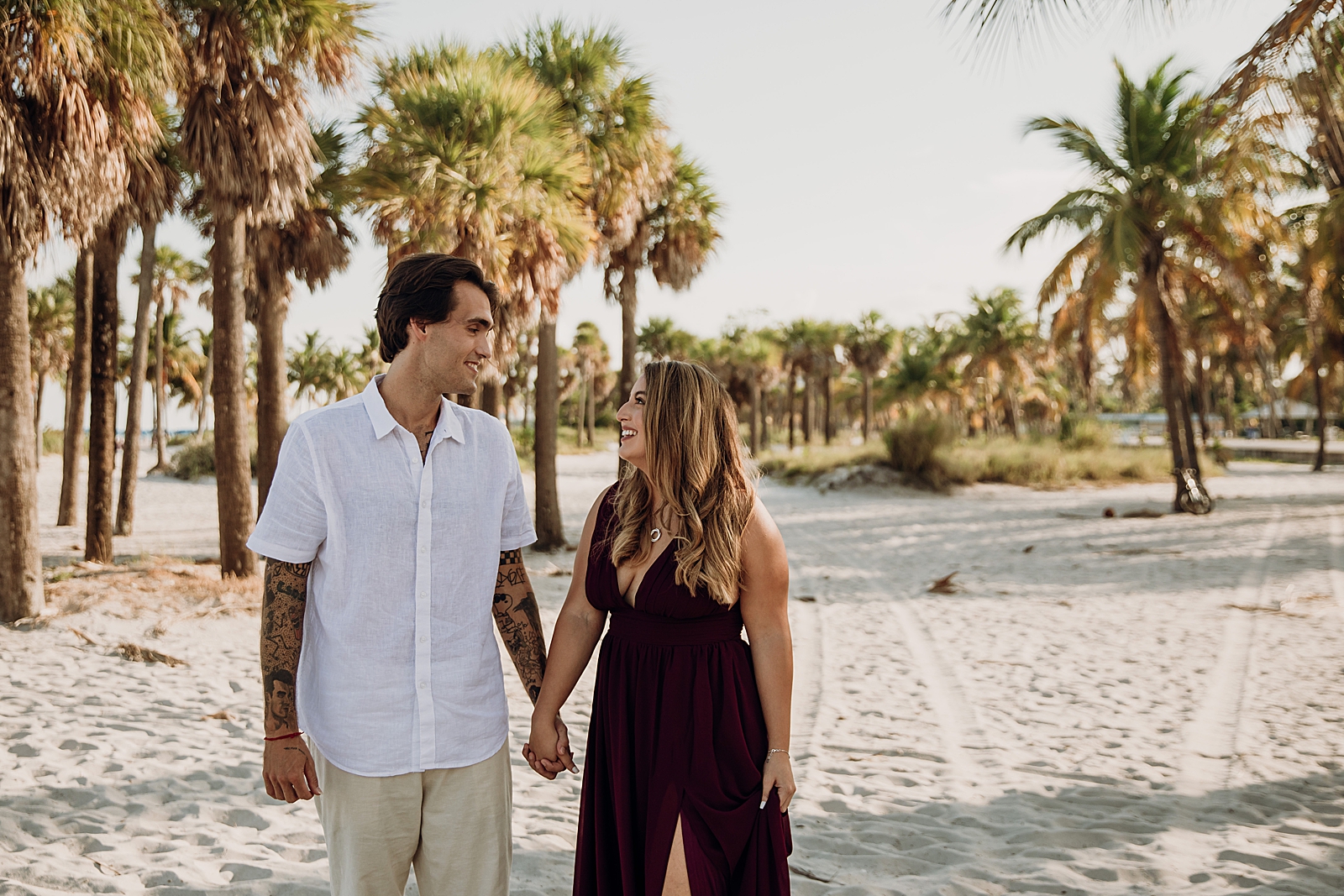 Couple holding hands and looking at each other on the sand with palm trees Key Biscayne Beach Engagement Photography captured by South Florida Family Photographer Maggie Alvarez Photography