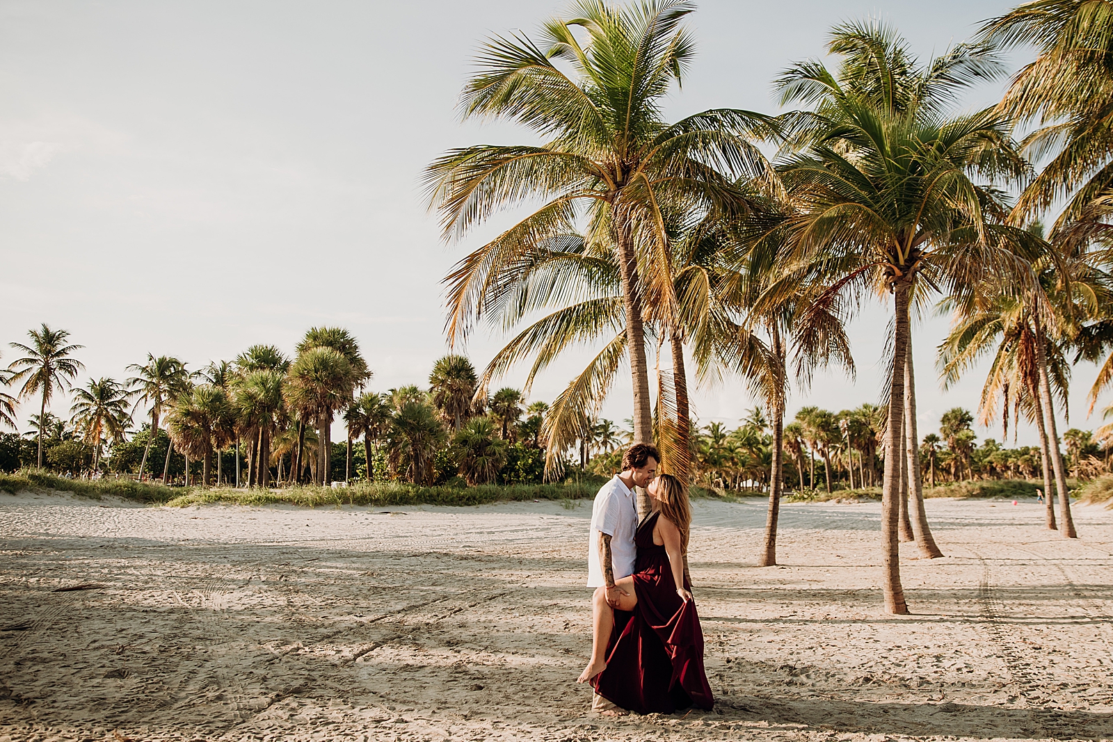 Wide shot off man lifting woman's leg and kissing her on the beach Key Biscayne Beach Engagement Photography captured by South Florida Family Photographer Maggie Alvarez Photography