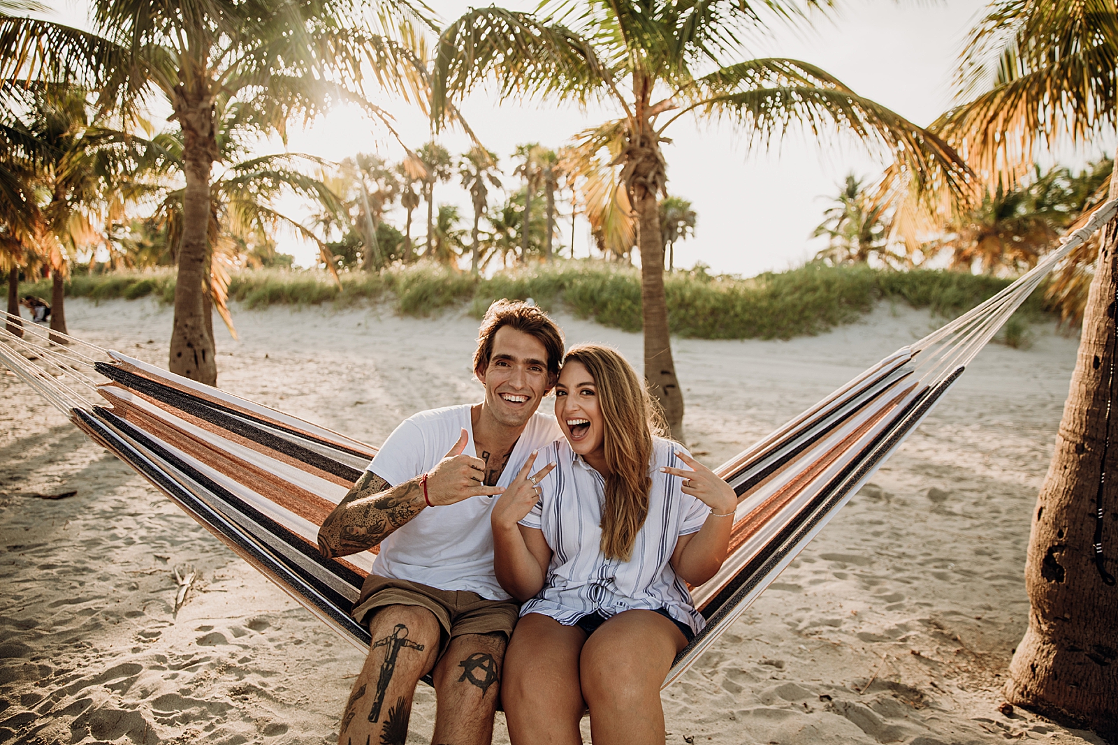 Couple hand signaling sitting on hammock on the beach Key Biscayne Beach Engagement Photography captured by South Florida Family Photographer Maggie Alvarez Photography