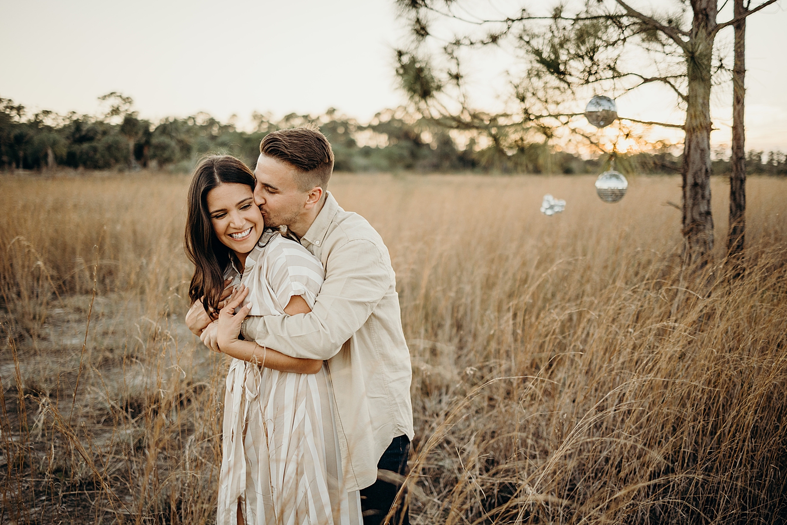 Man wrapping arms around woman with disco balls hanging on tree Royal Palm Nature Preserve Engagement Photography captured by South Florida Engagement Photographer Maggie Alvarez Photography