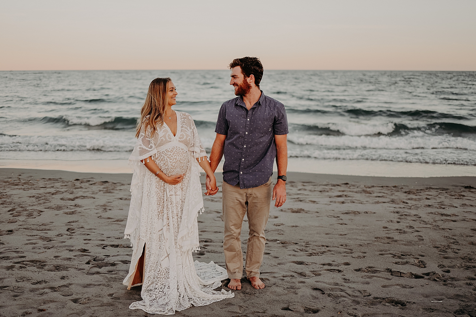 Pregnant wife holding baby bump and husband's hand in front of ocean water Spanish River Park Maternity Photography captured by South Florida Family Photographer Maggie Alvarez Photography