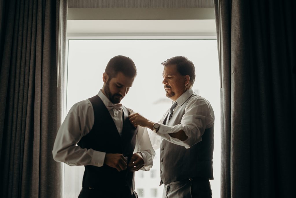 Groom putting vest by the window and Groomsmen assisting putting pocket square in