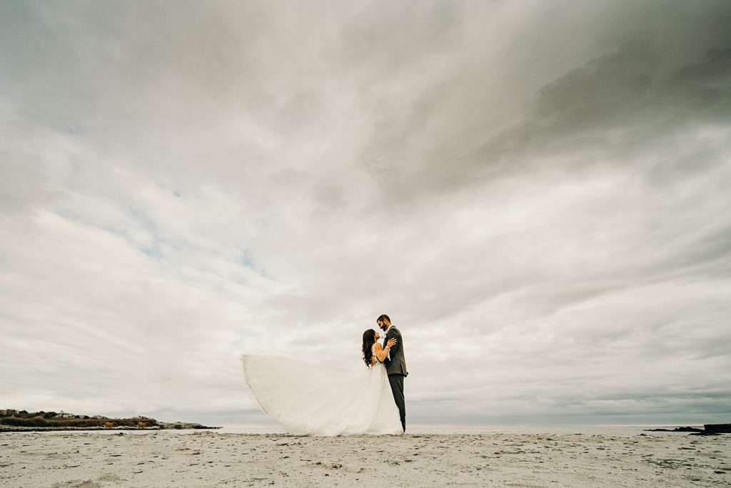 Wide below shot of Bride and Groom looking and holding each other on the sand with dress flowing