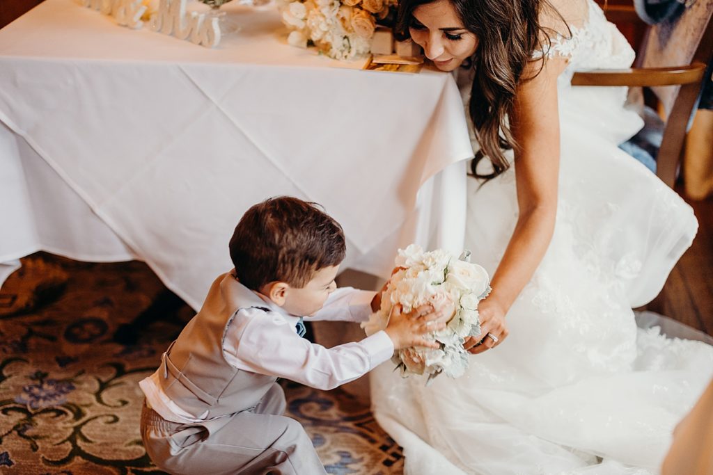 Bride sitting and handing bouquet to little boy by sweetheart table