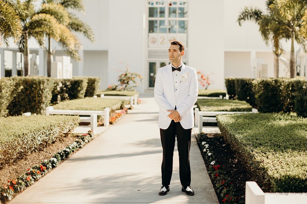 Groom in black and white suit standing on pathway by greenery