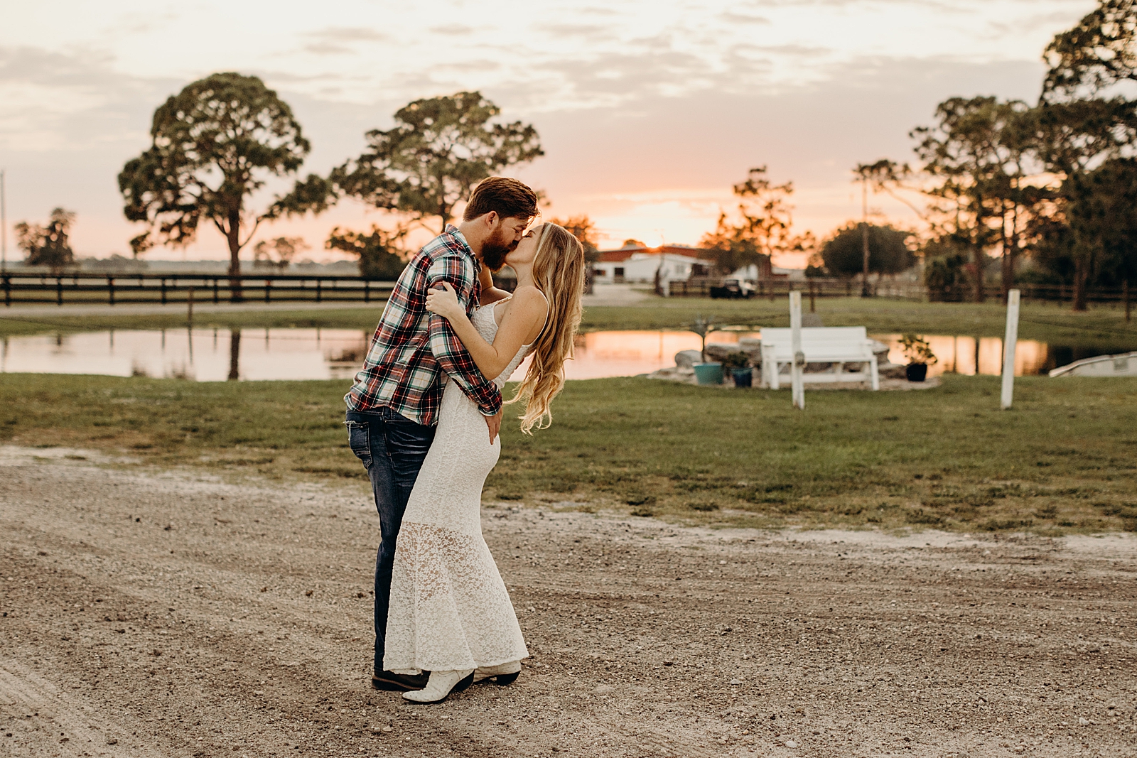 Couple holding each other close and kissing on dirt road on farm with the sunset behind them