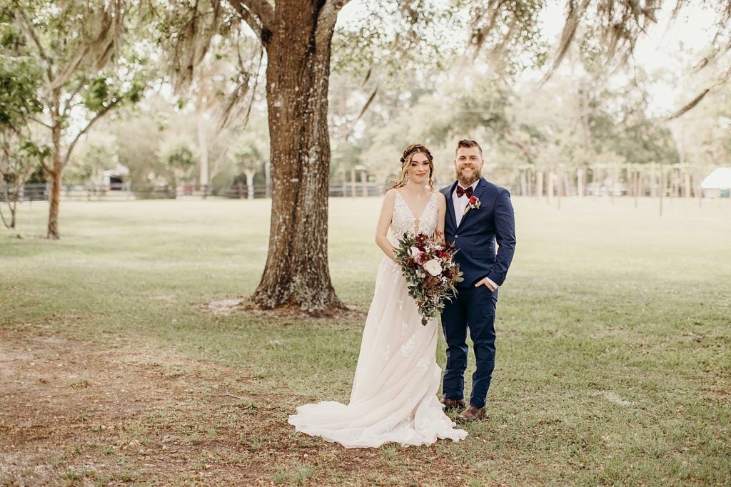 Bride and Groom standing together with extravagant bouquet by tree on green field 