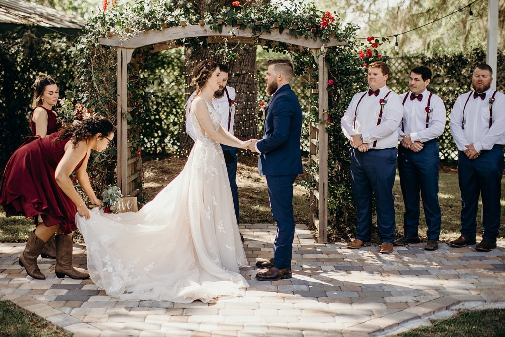 Bride and Groom hand in hand during Ceremony with Bridesmaid helping adjust dress