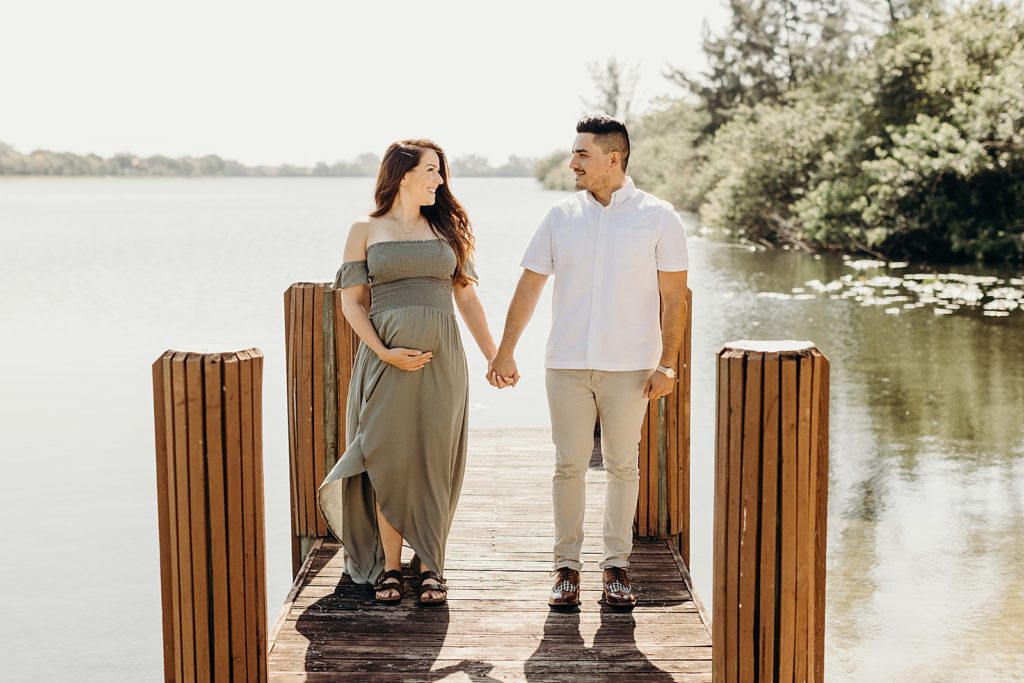 Couple holding hands and standing on small wooden dock with pregnant soon-to-be mother holding baby bump