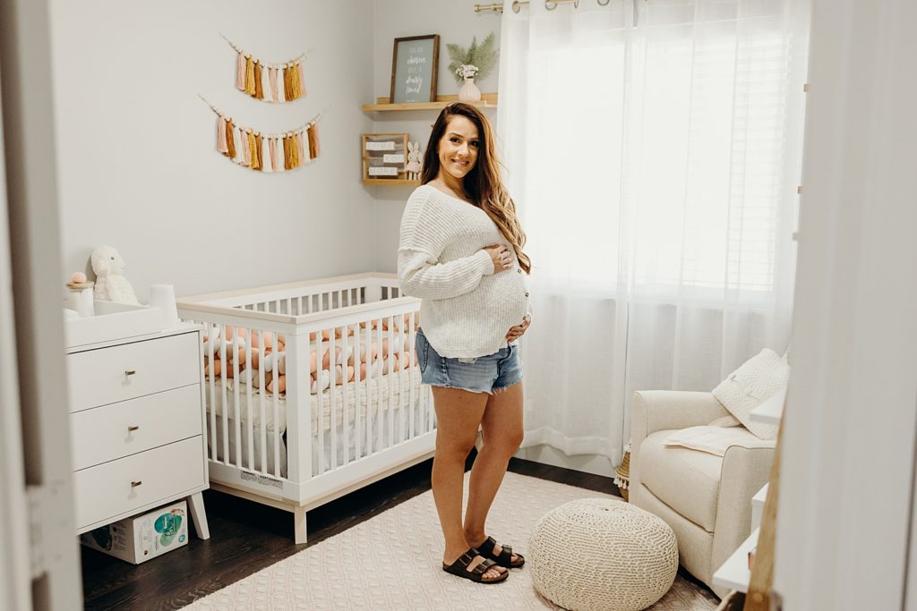 Pregnant Mother standing in bright Baby room with cradle and knitted decor