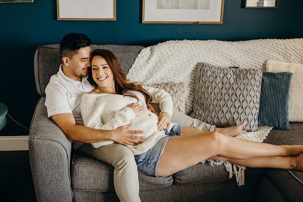 Pregnant woman laying on man sitting on grey couch while they holding her stomach