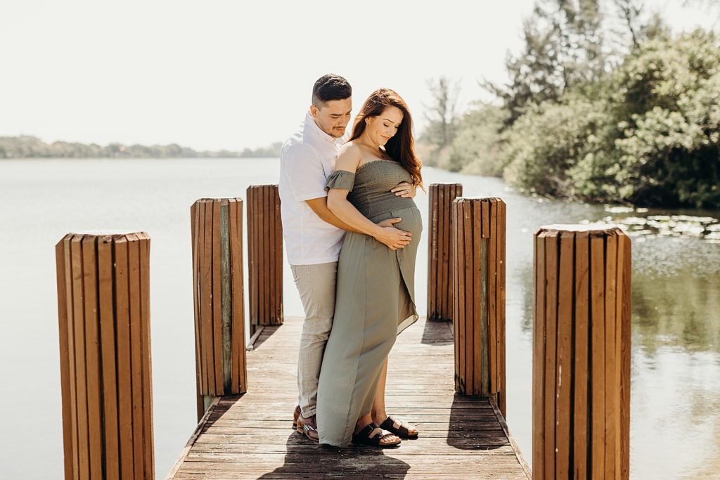 Husband holding pregnant wife's stomach from behind on small wooden dock on lake water
