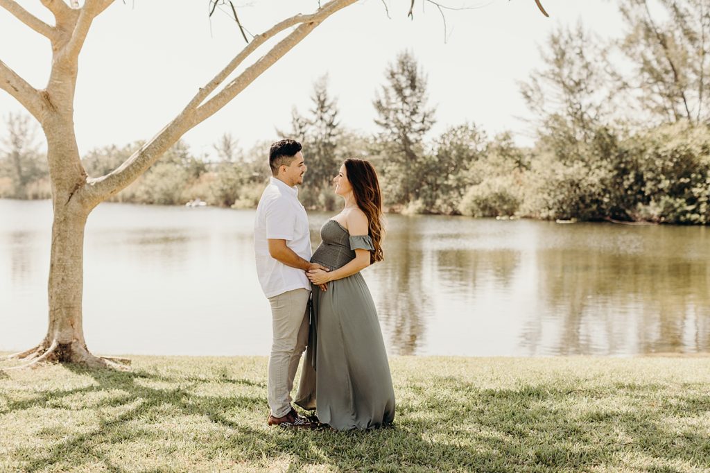 Pregnant couple standing face to face holding baby stomach next to calm lake