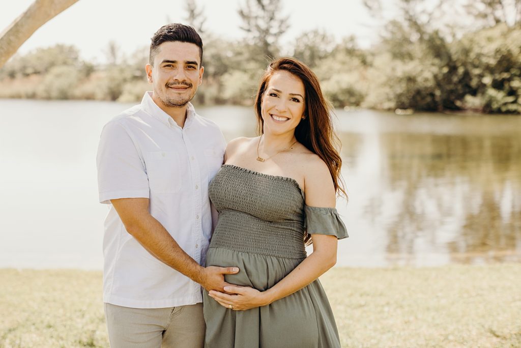 Couple standing next to each other with one hand each on pregnant stomach in front of calm lake