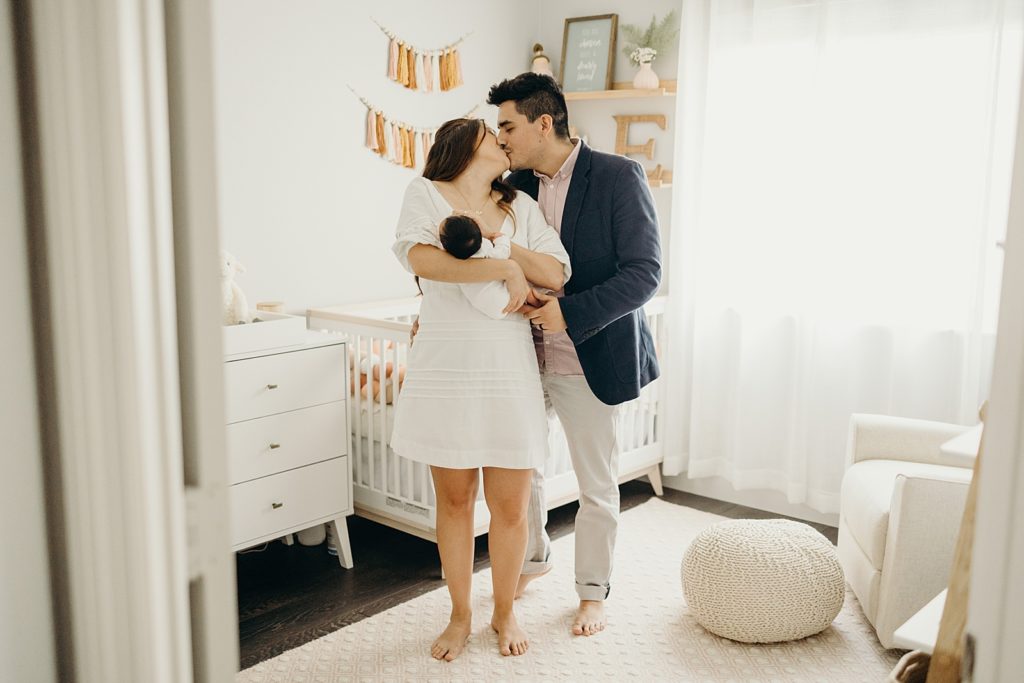 Couple holding each other and kissing as Mother holds their baby in baby room
