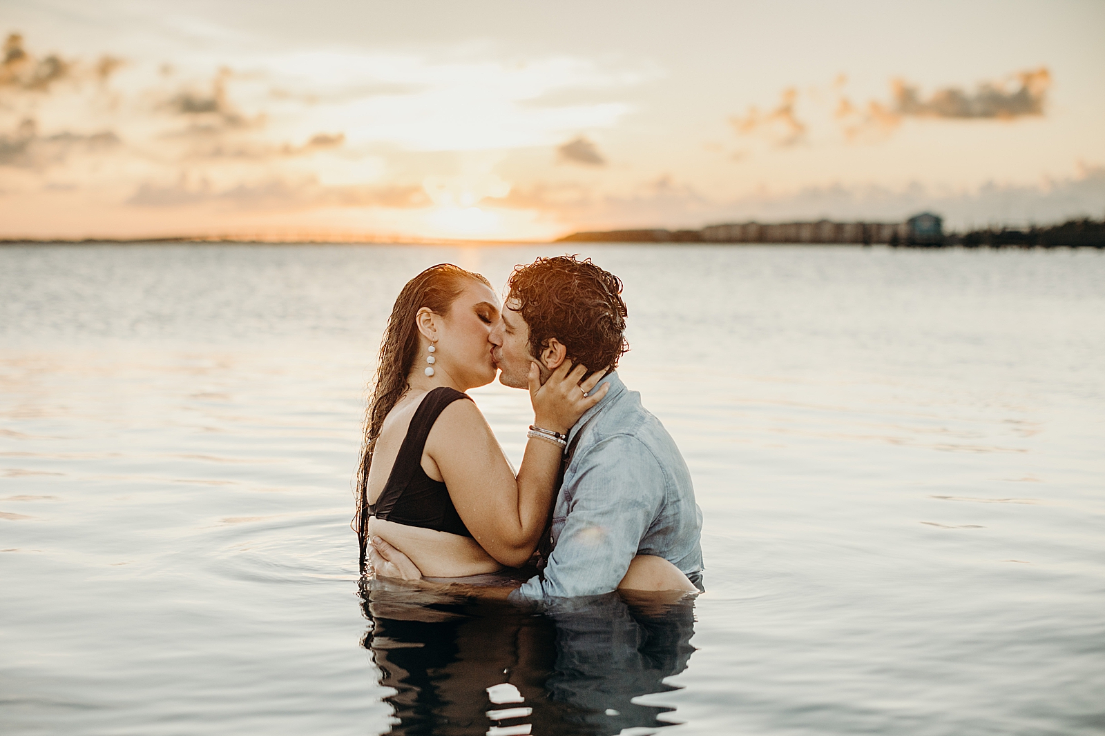 Couple holding each other and kissing in calm ocean water with sun setting