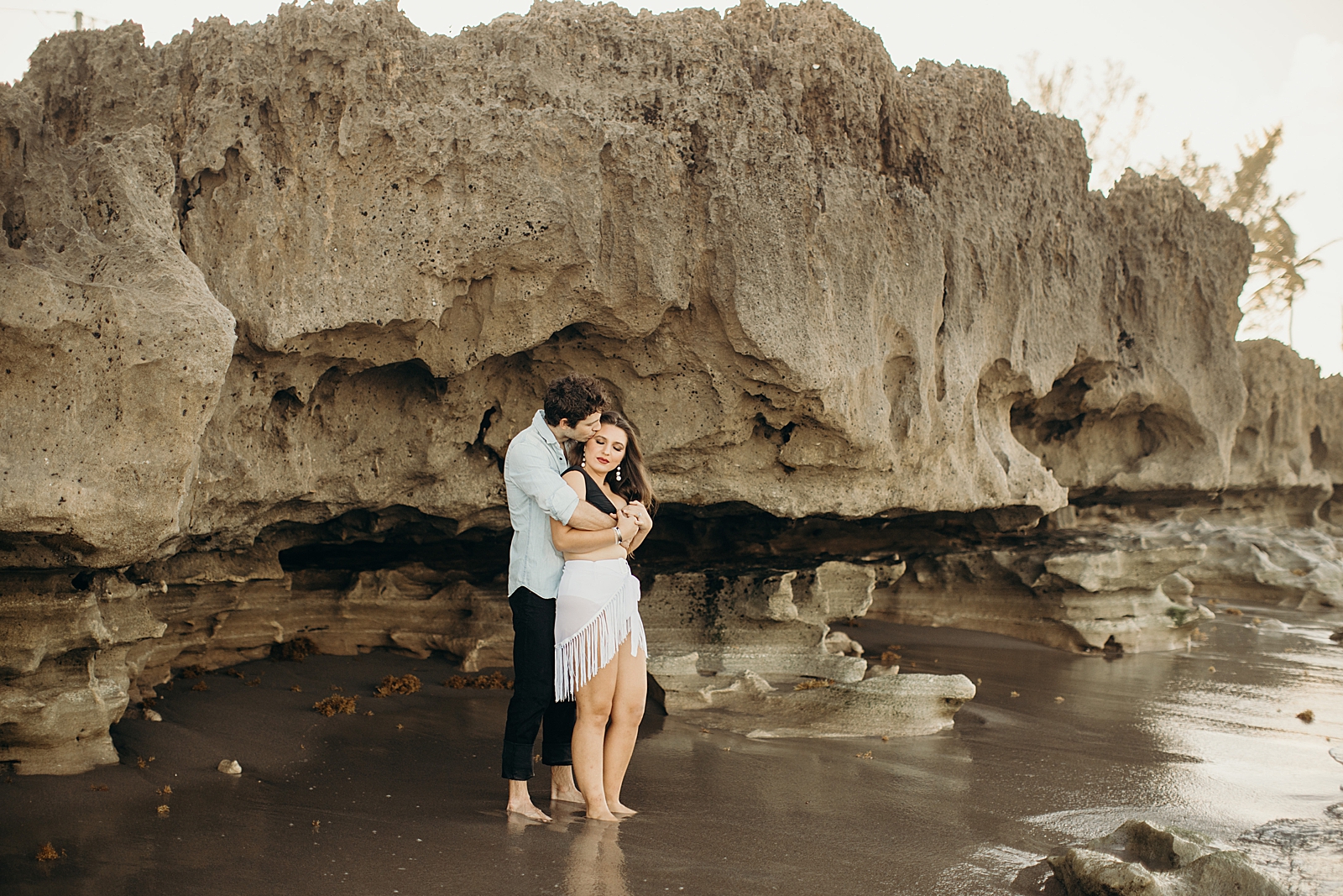Man holding woman from behind and kissing side of her forehead next to naturally rock formation by the wet sand