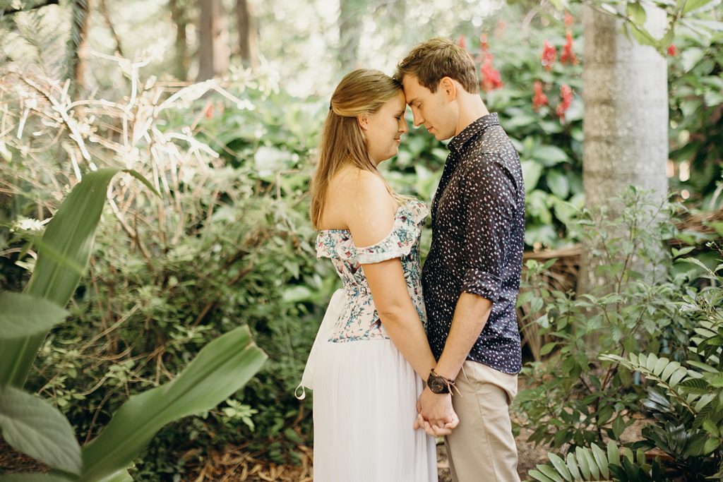 Couple holding hands and resting their foreheads on each other while standing in greenery exhibit