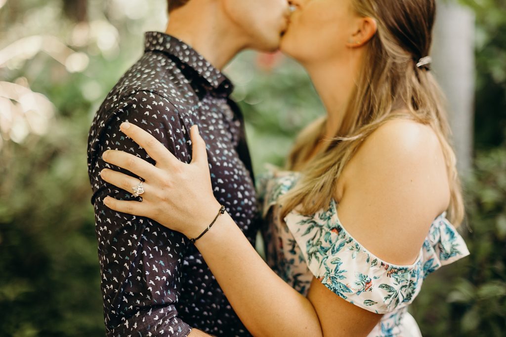 Closeup of couple standing in front of each other with woman's engagement ring hand on man's shoulder while couple kisses