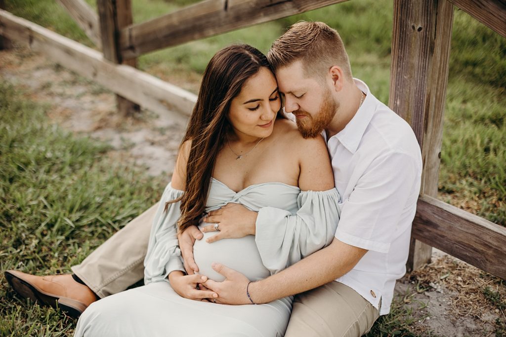 Couple sitting on ground leaning on wooden fence and holding pregnant stomach