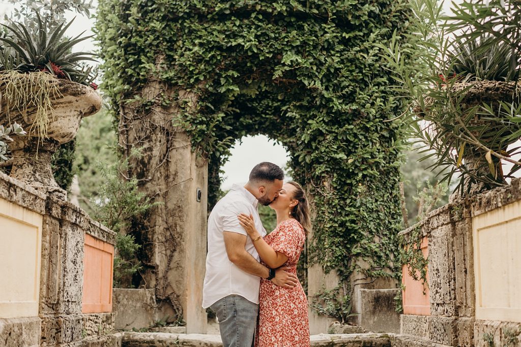 Couple holding and kissing each other in front of stone arch with greenery