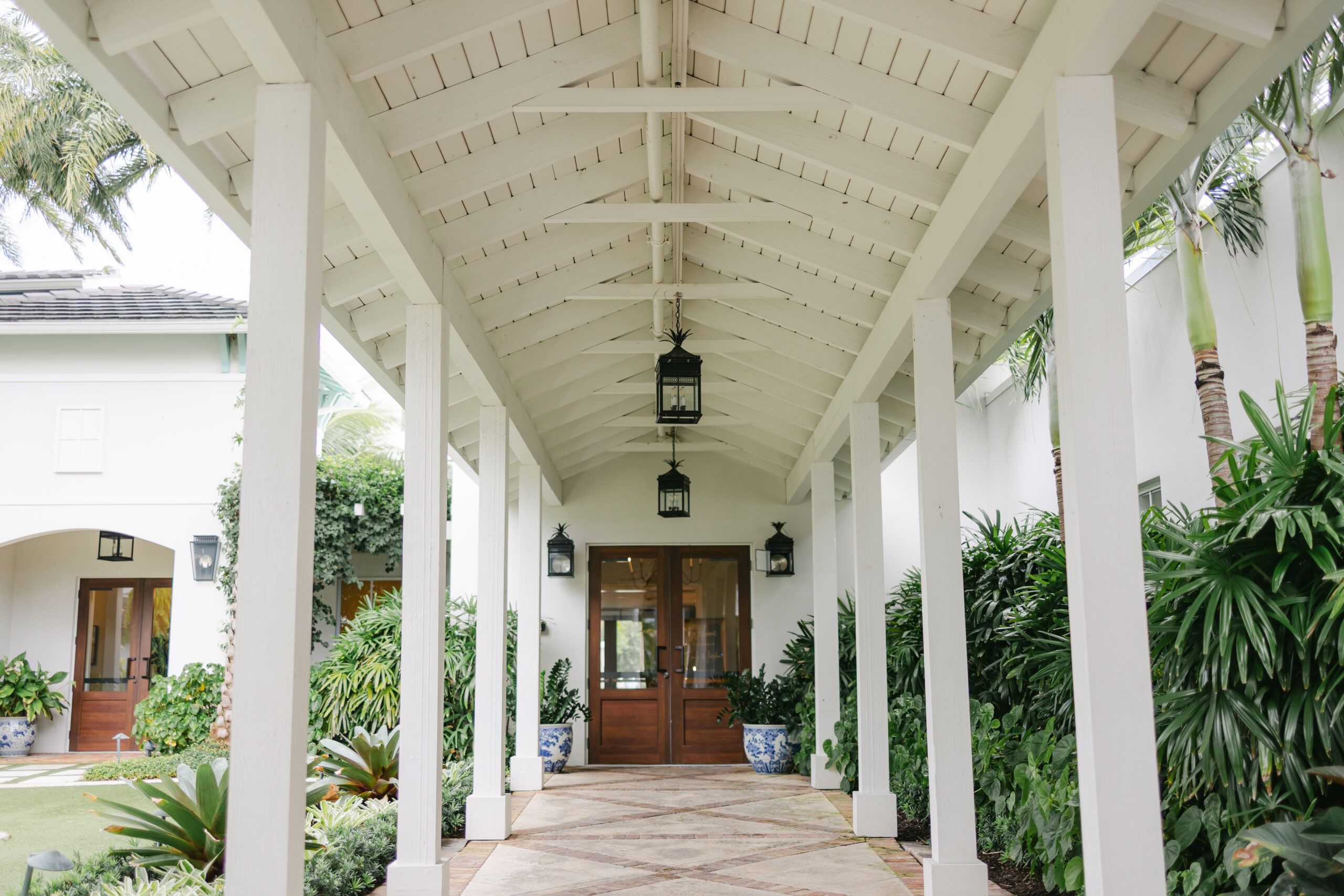 Covered walkway at the Pelican Club in Jupiter, Florida, featuring white columns, lantern lighting, lush tropical greenery, and elegant double doors—an ideal spot for wedding portraits.