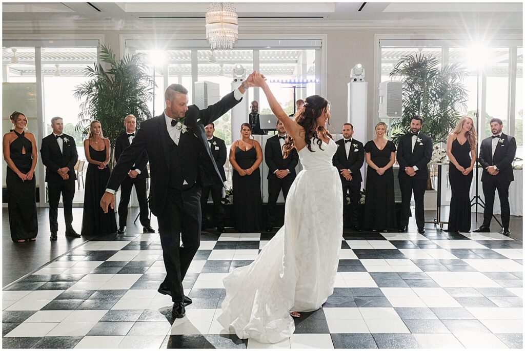 Bride and groom share their first dance at the Pelican Club in Jupiter, Florida, surrounded by their wedding party in formal black attire on a black-and-white checkered dance floor beneath elegant chandeliers.